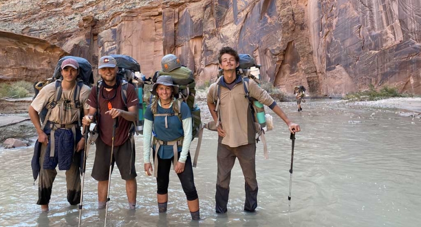 a group of gap year students wearing backpacks stand in a canyon in ankle-deep water